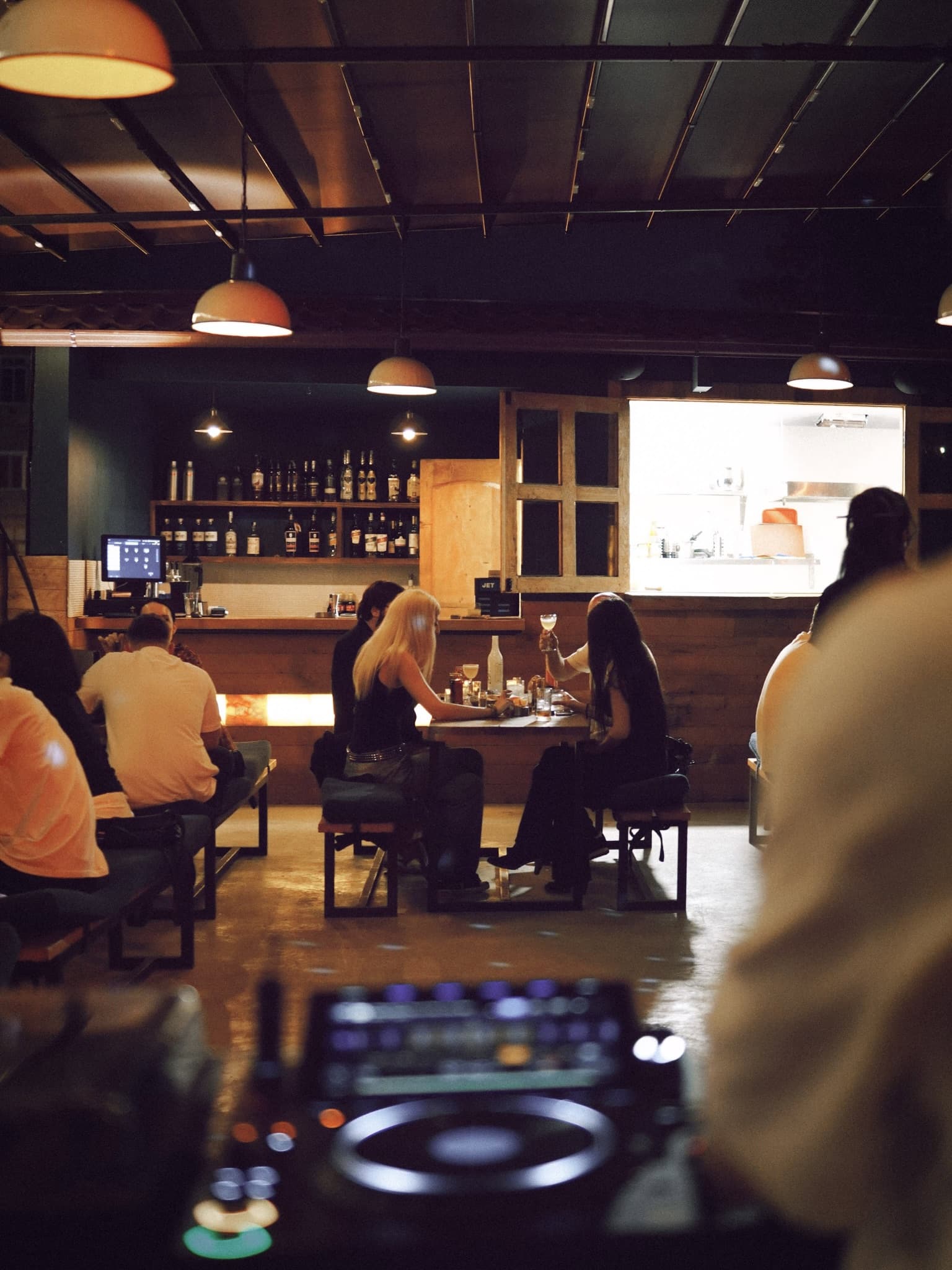 The bar under pendant lamps: guests at a long table, lit bottle shelves behind the wooden counter, a DJ deck in the foreground