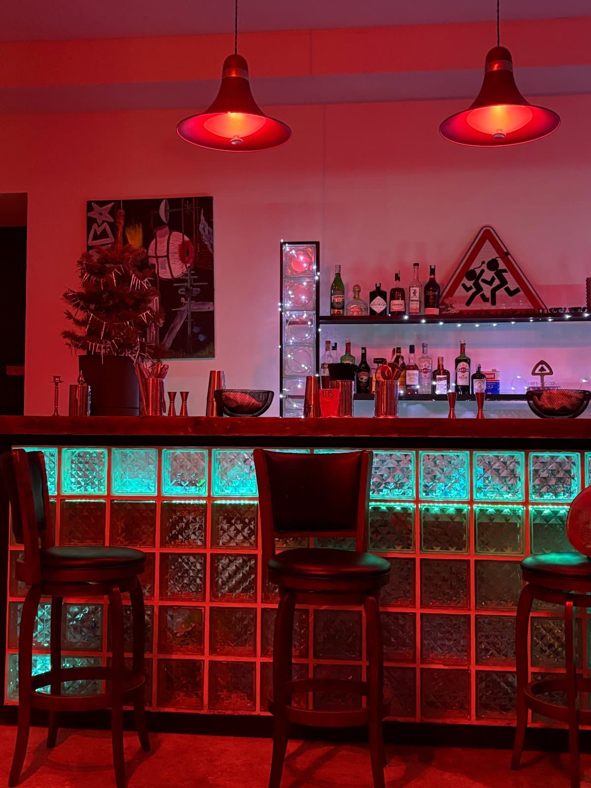 The bar front built from glass blocks lit teal and red, leather stools in front, two red pendant lamps and a lit bottle shelf behind